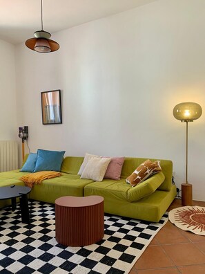 A living room with a green sofa, a round wooden coffee table, a black and white checkered rug, a floor lamp, and a framed picture on the wall.