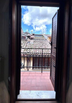 Vista a través de una ventana abierta con baranda de metal, que da a un tejado de baldosas y un edificio con fachada amarilla.
