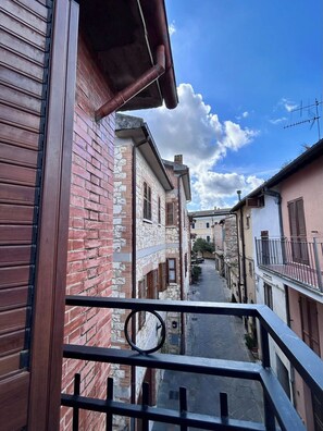 Una calle estrecha flanqueada por edificios, cielo despejado y barandilla de balcón.