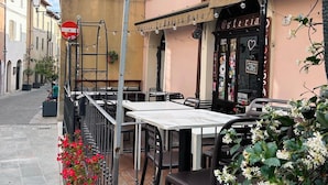 A street view with a building featuring outdoor seating, a sign reading 'Osteria,' and a flag.