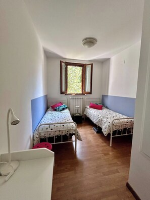 Habitación estrecha con dos camas individuales, una ventana, un radiador y una lámpara blanca.