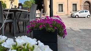 An outdoor seating area with wicker chairs and a table, a planter with white and pink flowers, and a building with arched windows in the background.