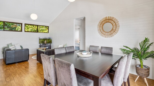 A modern living room with a dining area, featuring a large dark wooden table, grey upholstered chairs, a grey sofa, and a television. - Quaint 2-bed Beach Abode With Ocean Views (Sunshine Coast)