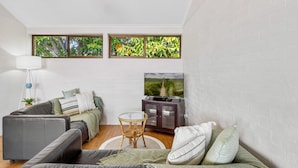 Una sala de estar moderna con un sofá gris, una mesita redonda y un televisor sobre un mueble bajo. Se ven ventanas con vistas a zonas verdes.
