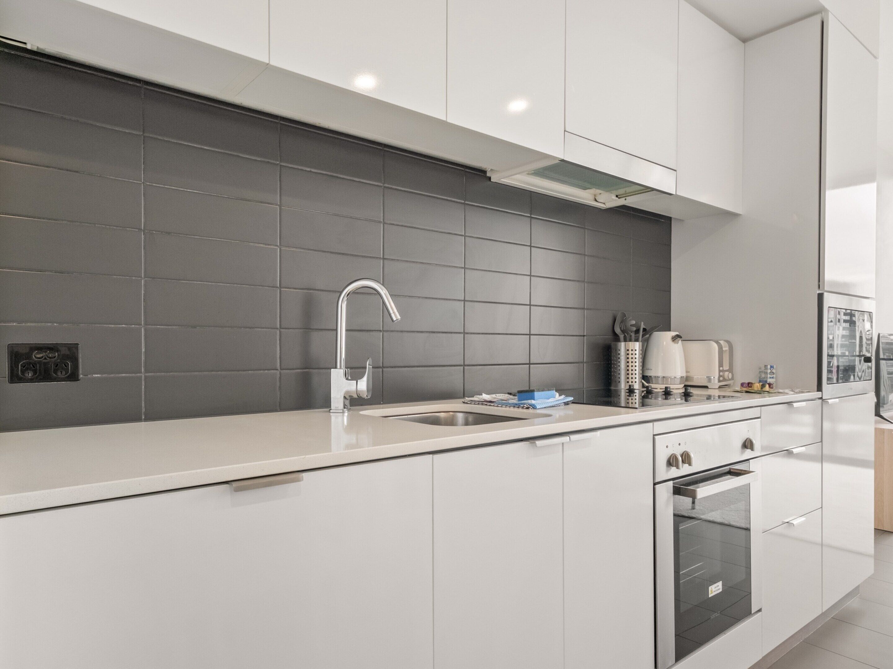 A modern kitchen with white cabinets, a grey backsplash, a stainless steel sink, and a built-in oven.