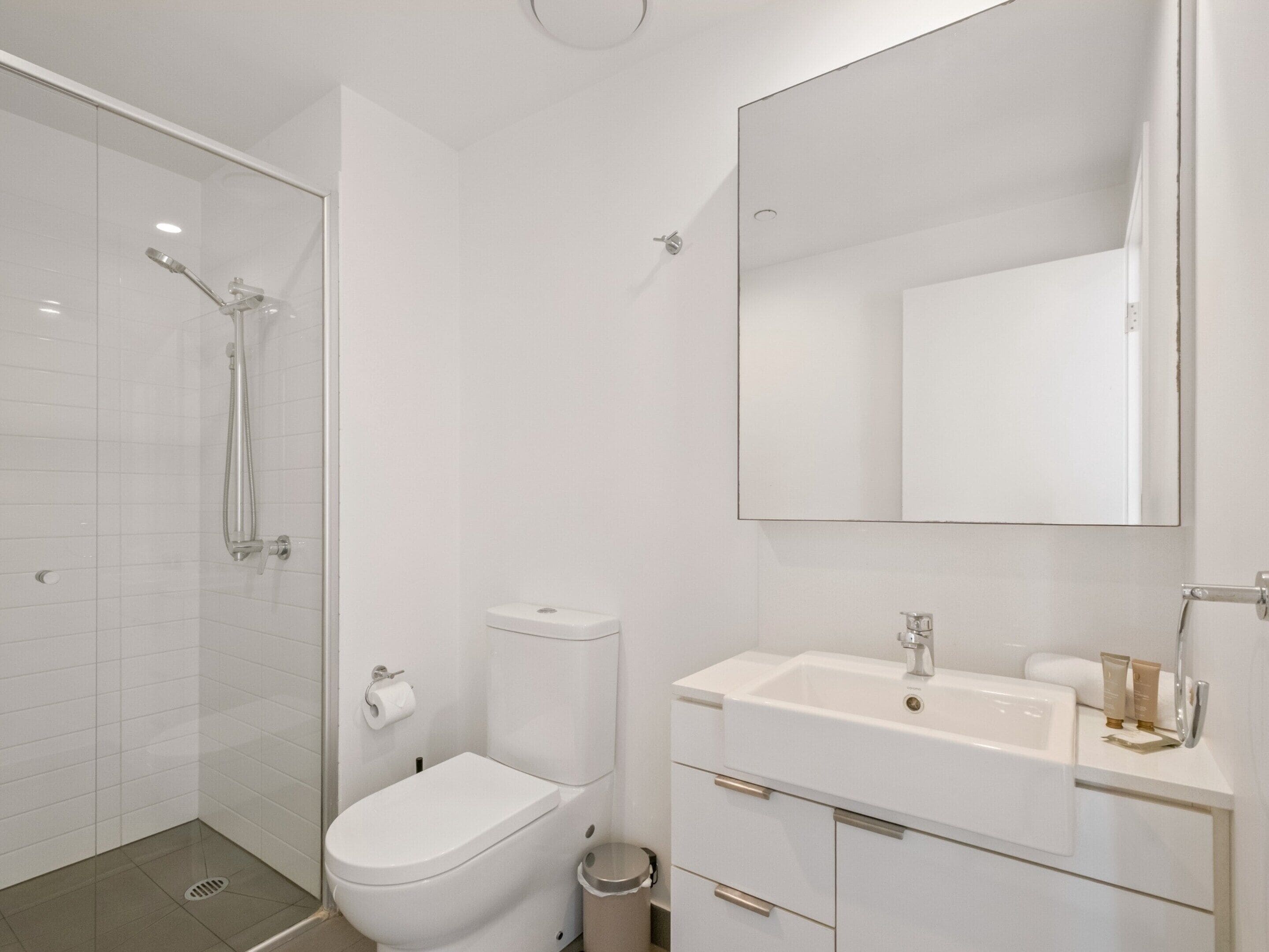 A modern bathroom with a glass shower enclosure, a toilet, a sink with a mirror, and a trash bin.