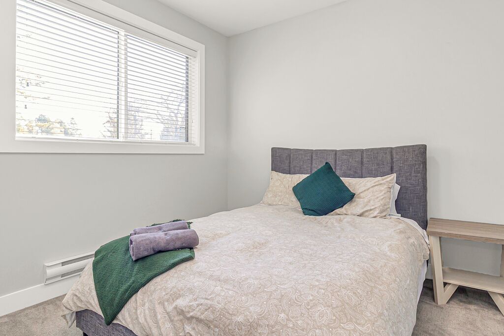 A bedroom with a bed, a window with blinds, a bedside table, and a view of the outside.