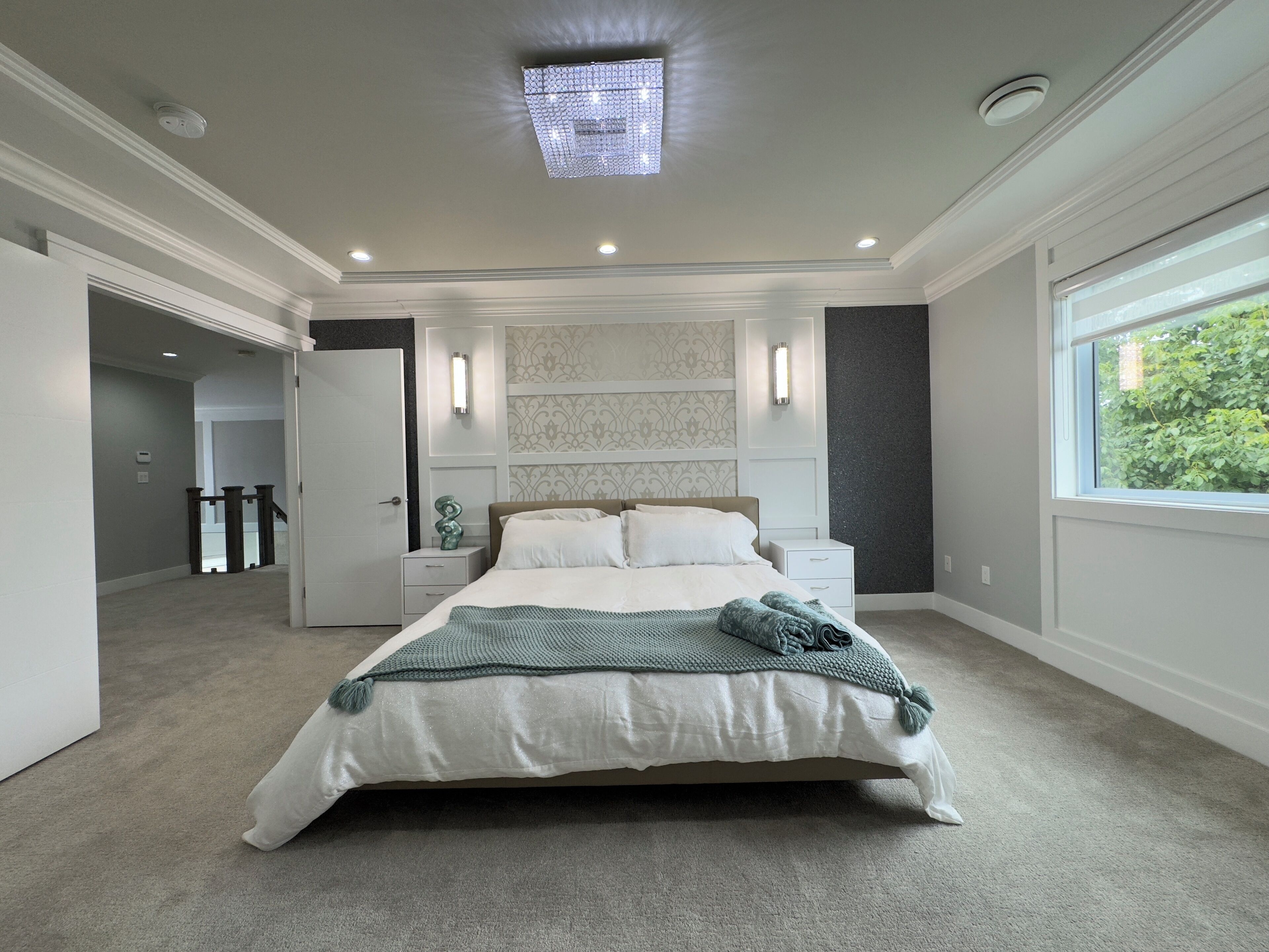 A modern bedroom with a large bed, bedside tables, a nightstand, a decorative wall panel, and a window with a view of greenery.
