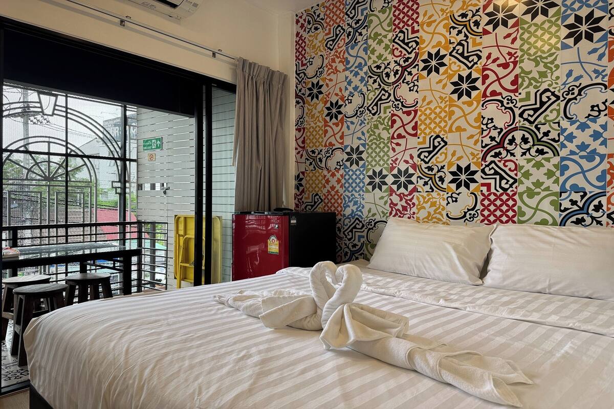 A hotel room with a bed, a window with a view, a patterned wall, and a small table with stools.