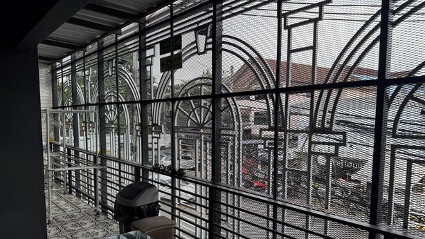 A view through a metal grid window with a view of a street and a building with large circular structures. - Phukhao Hostel (Chiang Mai)