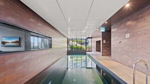 A modern indoor swimming pool with clear glass walls, a tiled floor, and a view of greenery outside. - Modern 1-bed City Apartment With Pool and Gym (Melbourne)