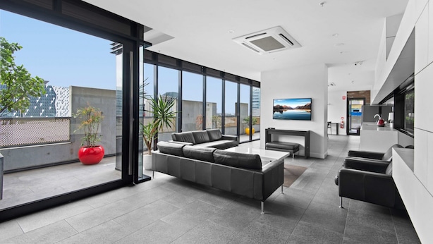 A modern living room with a sectional sofa, a flat-screen TV, and a large window offering a city view. - Modern 1-bed City Apartment With Pool and Gym (Melbourne)