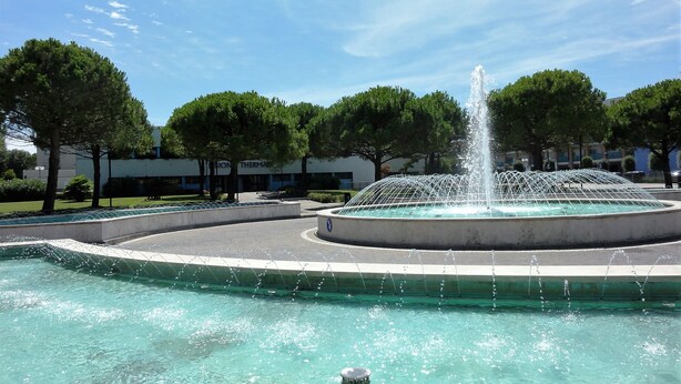A fountain with water jets in a circular pool, surrounded by trees and a building in the background. - SUN CHARIOTEER - C2 4+0 - A32168 (Bibione)
