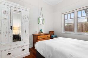 Ein Schlafzimmer mit einem weißen Bett, einem Holfnachtkästchen, einer Kommode mit Spiegel und einem Fenster mit Blick auf einen Zaun.