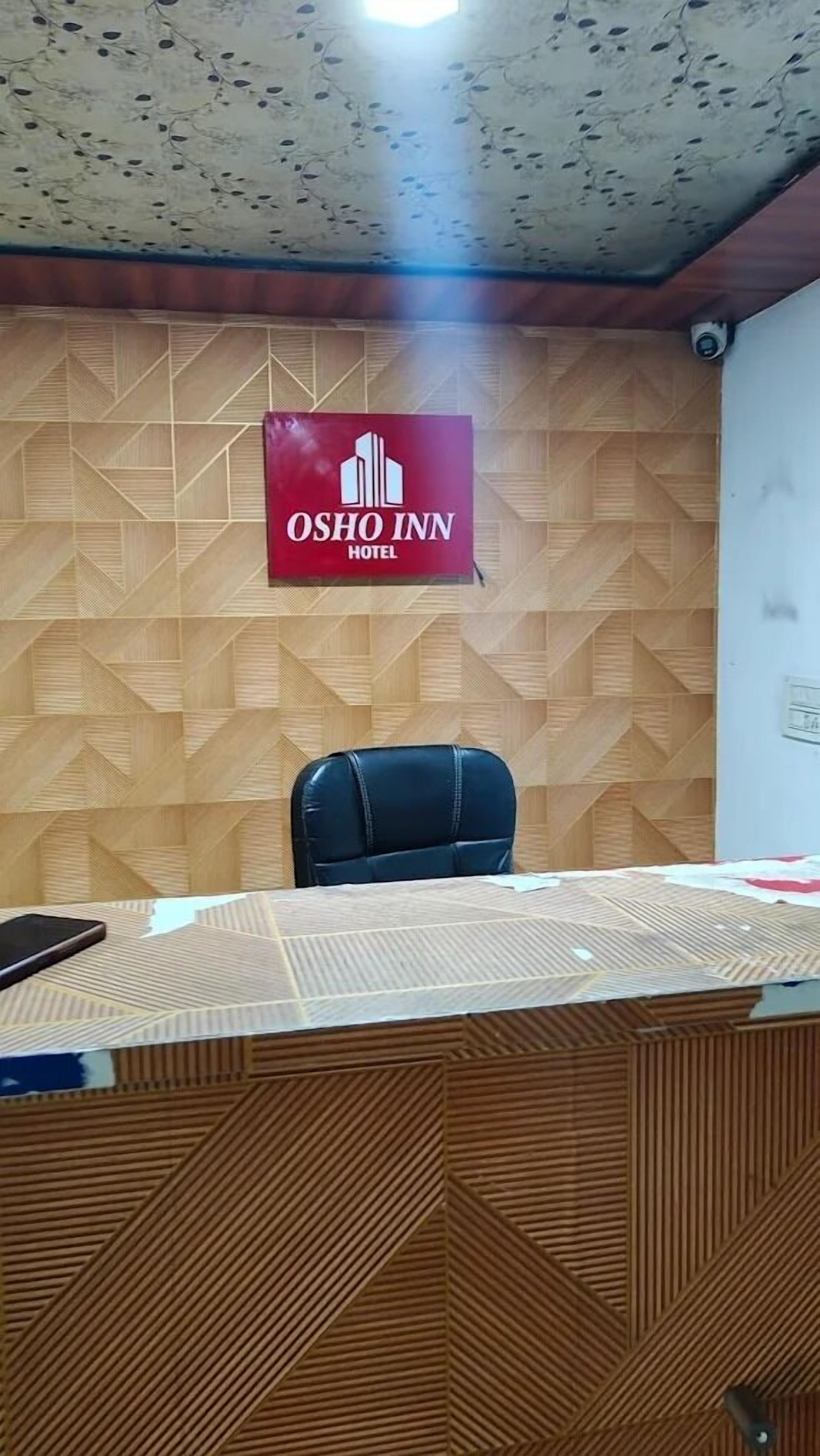 Una sala de conferencias con una pared de madera con diseño, una silla de oficina negra y una mesa cubierta con un mantel transparente. Se ve un letrero del Hotel Osho Inn en la pared.