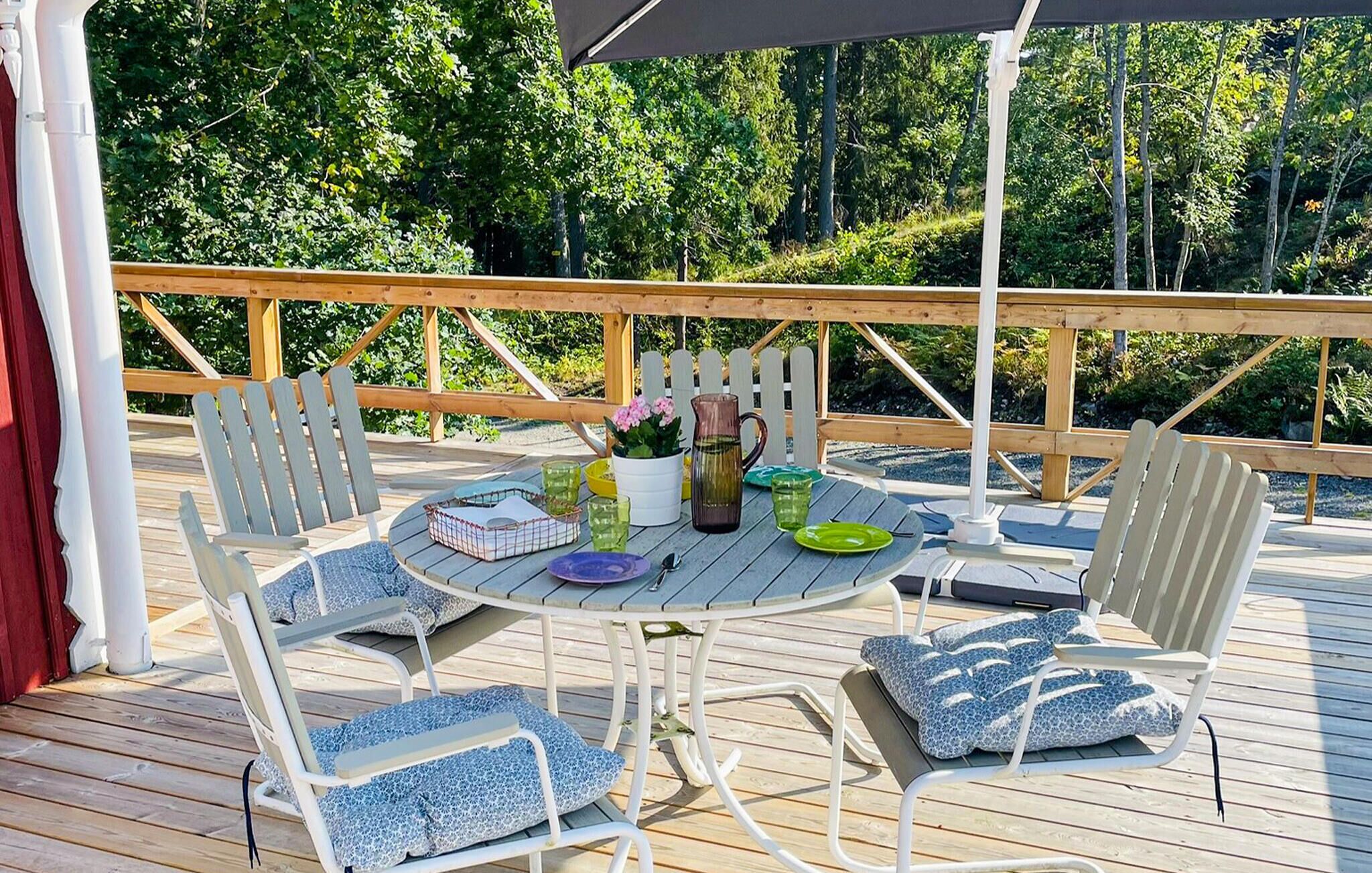 Una terraza de madera con una mesa redonda, sillas blancas y un paraguas negro.