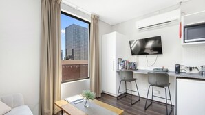 A modern hotel room with a large window, a flat-screen TV, a small kitchenette, and a seating area with a bench and two bar stools. - An Oasis in the City - Studio - by Sepal Stays (Perth)