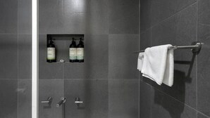 A modern bathroom with dark tiles, a glass-enclosed shower, a towel rack, and two bottles of shampoo. - An Oasis in the City - Studio - by Sepal Stays (Perth)