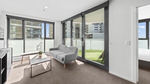 Una sala de estar moderna con un sofá gris, una mesa de centro y un amplio ventanal corredizo que da a un balcón con vistas a edificios y una zona verde.