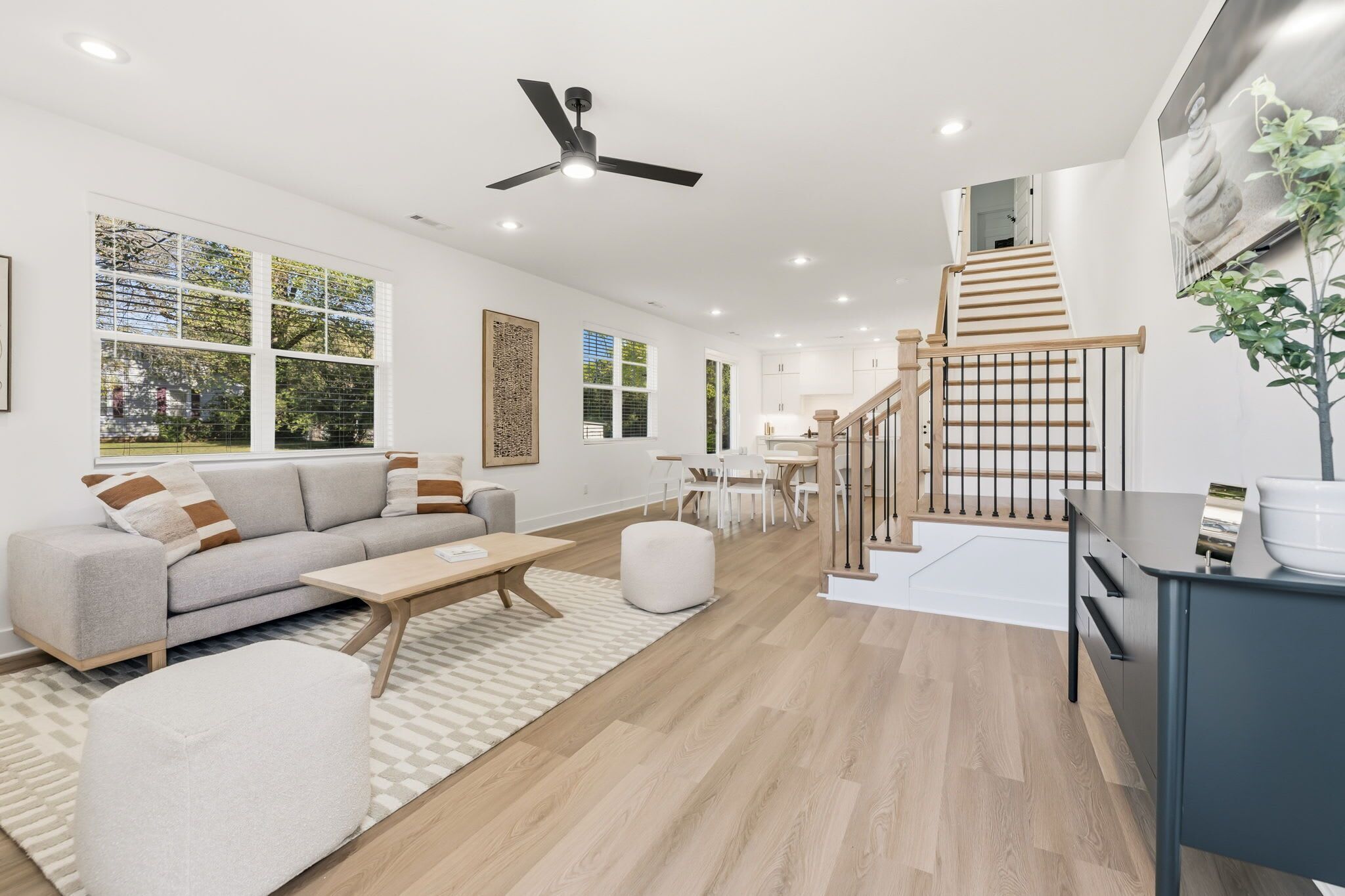 Una sala de estar moderna con sofá, mesa de centro y reposapiés, una escalera que lleva a una cocina, y ventanas amplias con persianas.