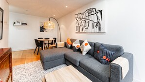Ein modernes Wohnzimmer mit einer grauen Eckcouch, einem Holztisch und einem Essbereich mit kleinem Tisch und Stühlen.