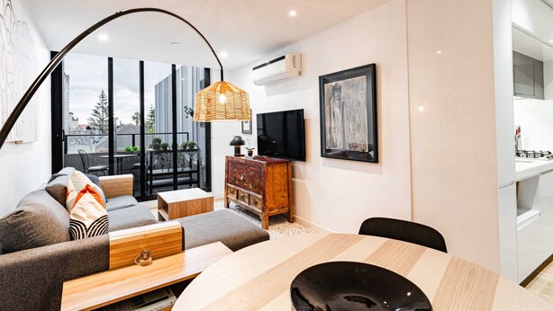 Ein modernes Wohnzimmer mit Sofa, Couchtisch und Esstisch. Ein Fernseher ist an der Wand befestigt und ein Fenster bietet Blick auf umliegende Gebäude.