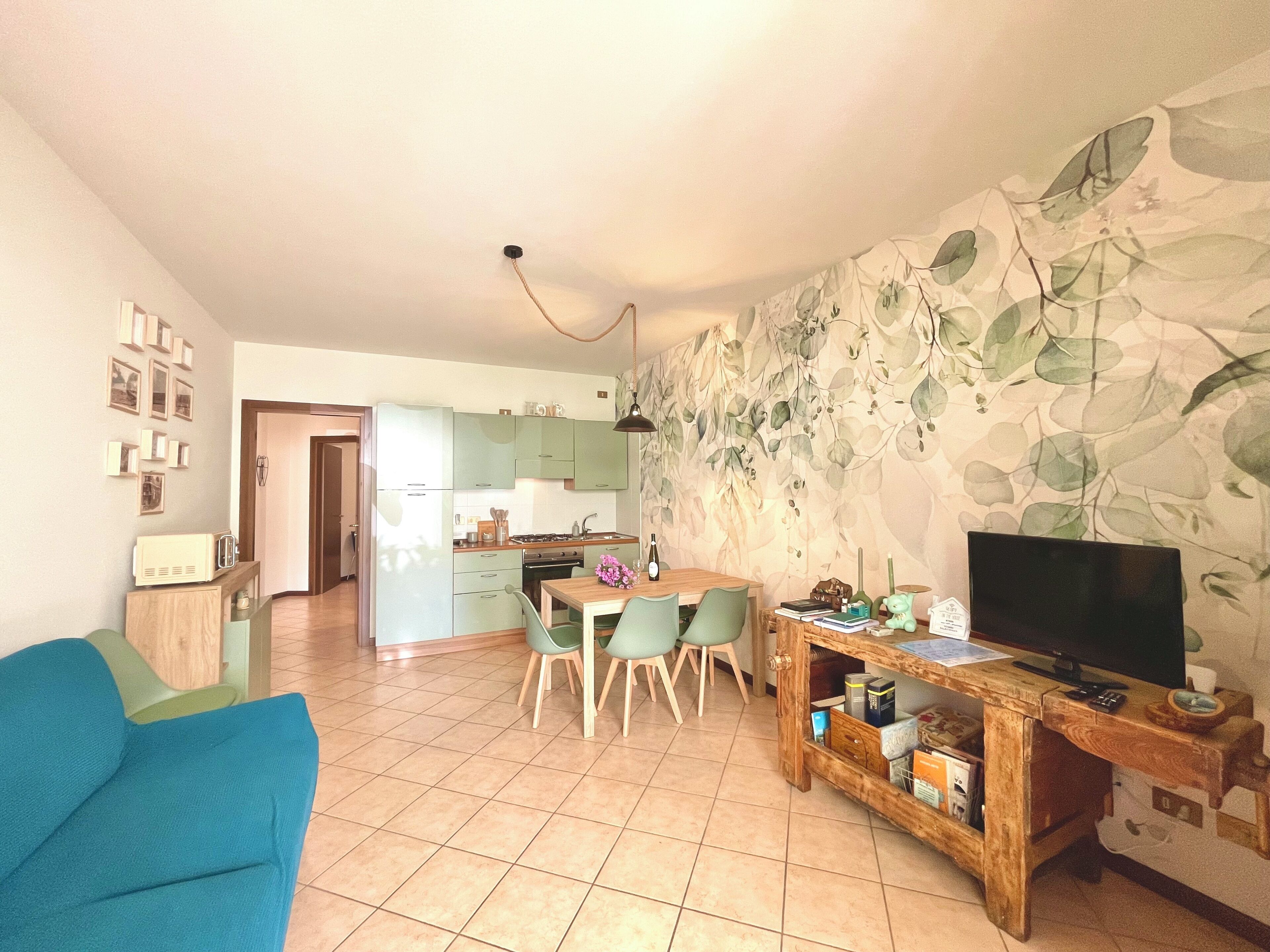A living room with a blue sofa, a wooden coffee table, and a TV on a wooden stand. There is a dining area with a table and chairs, and a kitchen in the background.
