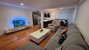 A modern living room with a flat-screen TV, a sofa, a coffee table, and a kitchen area in the background. - Northshore Central Cosy & Comfortable 3BR Home (Auckland)