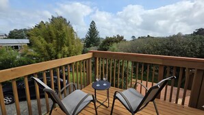 A wooden deck with two chairs and a small table, overlooking a garden and a residential area. - Northshore Central Cosy & Comfortable 3BR Home (Auckland)
