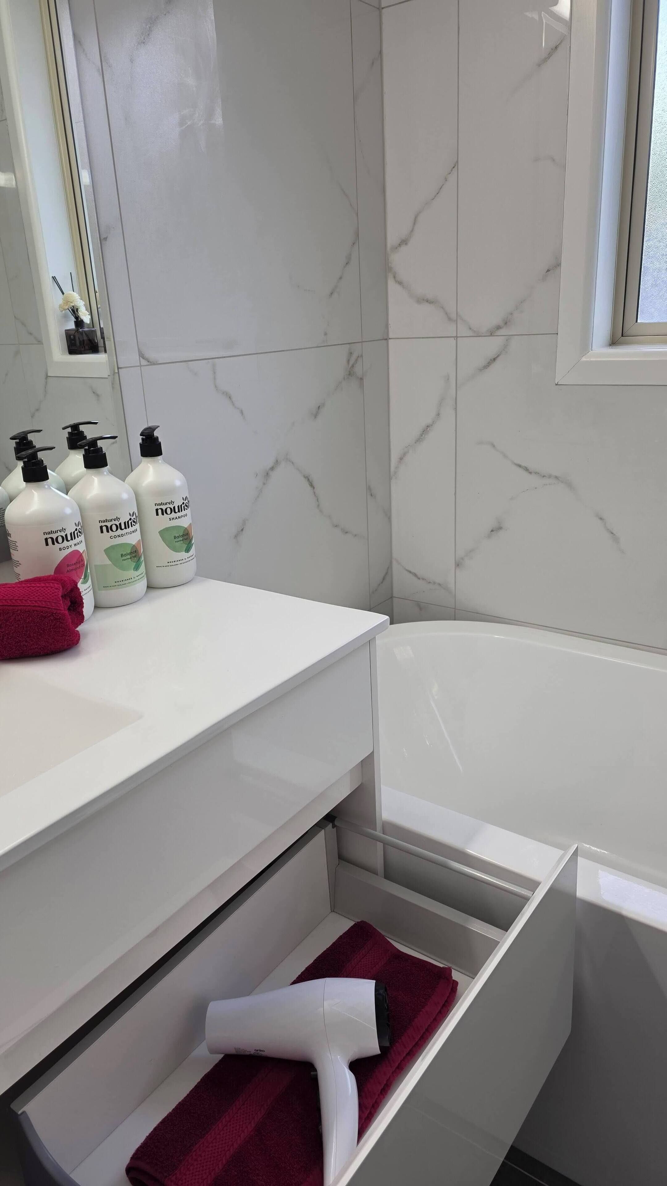 Une salle de bain avec un meuble-lavabo blanc, un miroir, une fenêtre et une baignoire. On y trouve trois flacons de gel douche et un sèche-cheveux posés sur le meuble-lavabo.