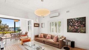Una sala de estar moderna con una mesa de centro de vidrio, un sofá marrón y un amplio ventanal con contraventanas.