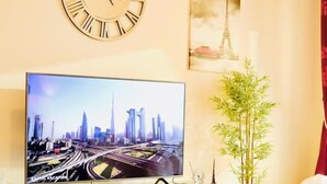 Una sala de estar con un televisor de pantalla plana grande mostrando un paisaje urbano, un reloj de pared redondo, un cuadro enmarcado de la Torre Eiffel, una planta de bambú y una mesa de centro moderna.