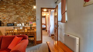 Una sala de estar acogedora con un sofá rojo, una mesa de comedor de madera y sillas. Se ve una zona de cocina con una pared de piedra y un radiador al fondo.