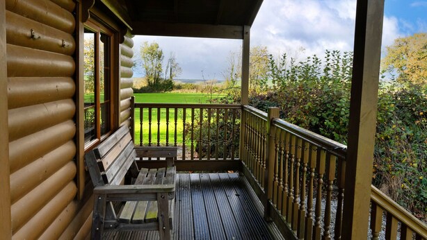 A wooden cabin with a porch, a bench, and a view of a green field. - Tarka's Holt Log Cabin, Stowford (Torrington)
