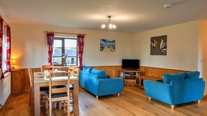 A living room with a wooden floor, a dining table with chairs, a blue sofa, a TV stand, and a window with curtains. - Tarka's Holt Log Cabin, Stowford (Torrington)
