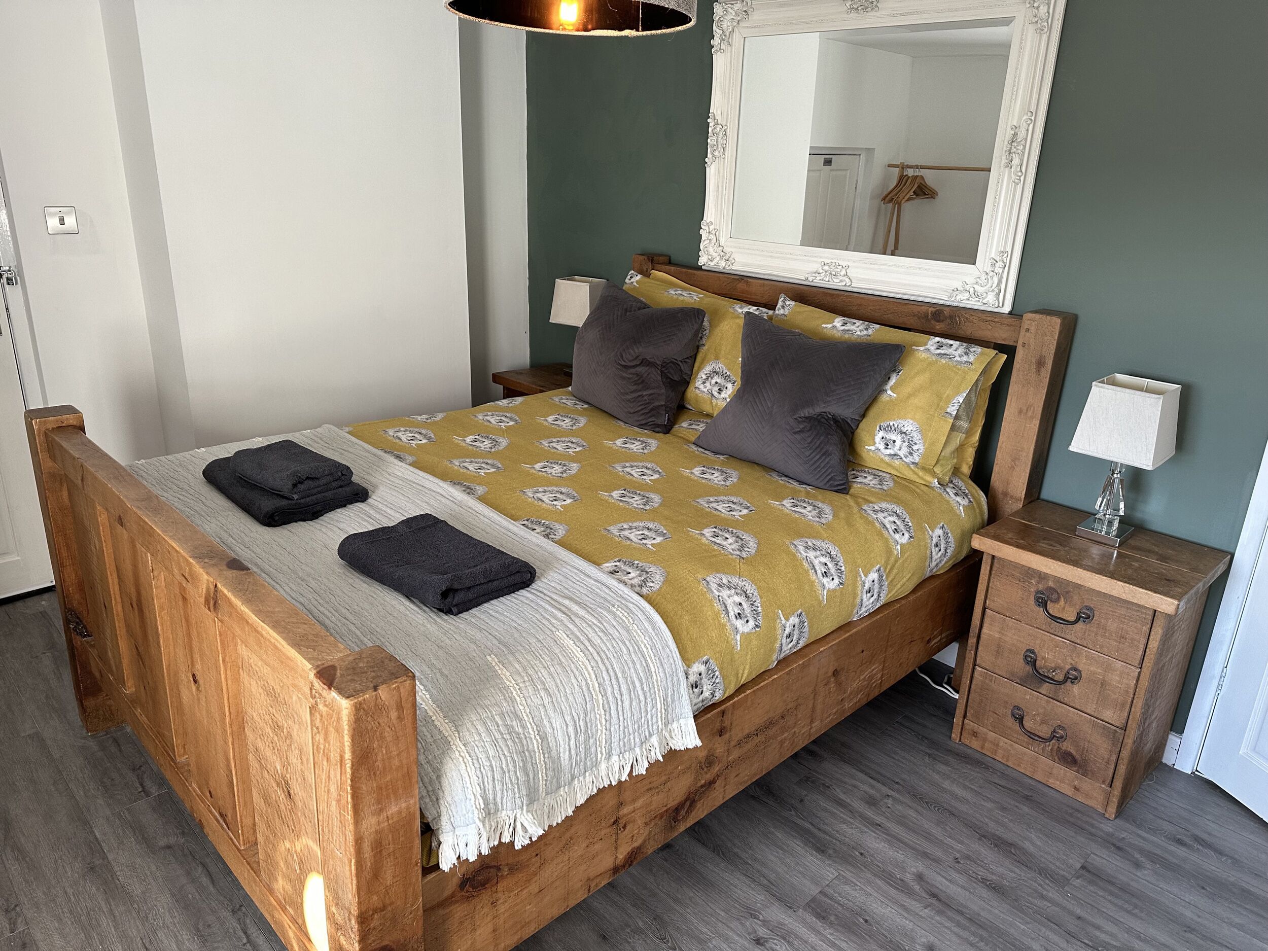 Un lit en bois avec une couvre-lit à motifs, deux oreillers, une table de chevet avec une lampe et un miroir au-dessus du lit.