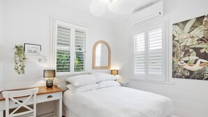 Une chambre à coucher avec un lit, un bureau et une chaise. Il y a une fenêtre avec des volets et une plante sur le bureau.