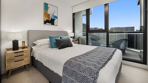 Un dormitorio moderno con una cama grande, una mesita de noche, una lámpara, un cuadro y un balcón con vistas a la ciudad.