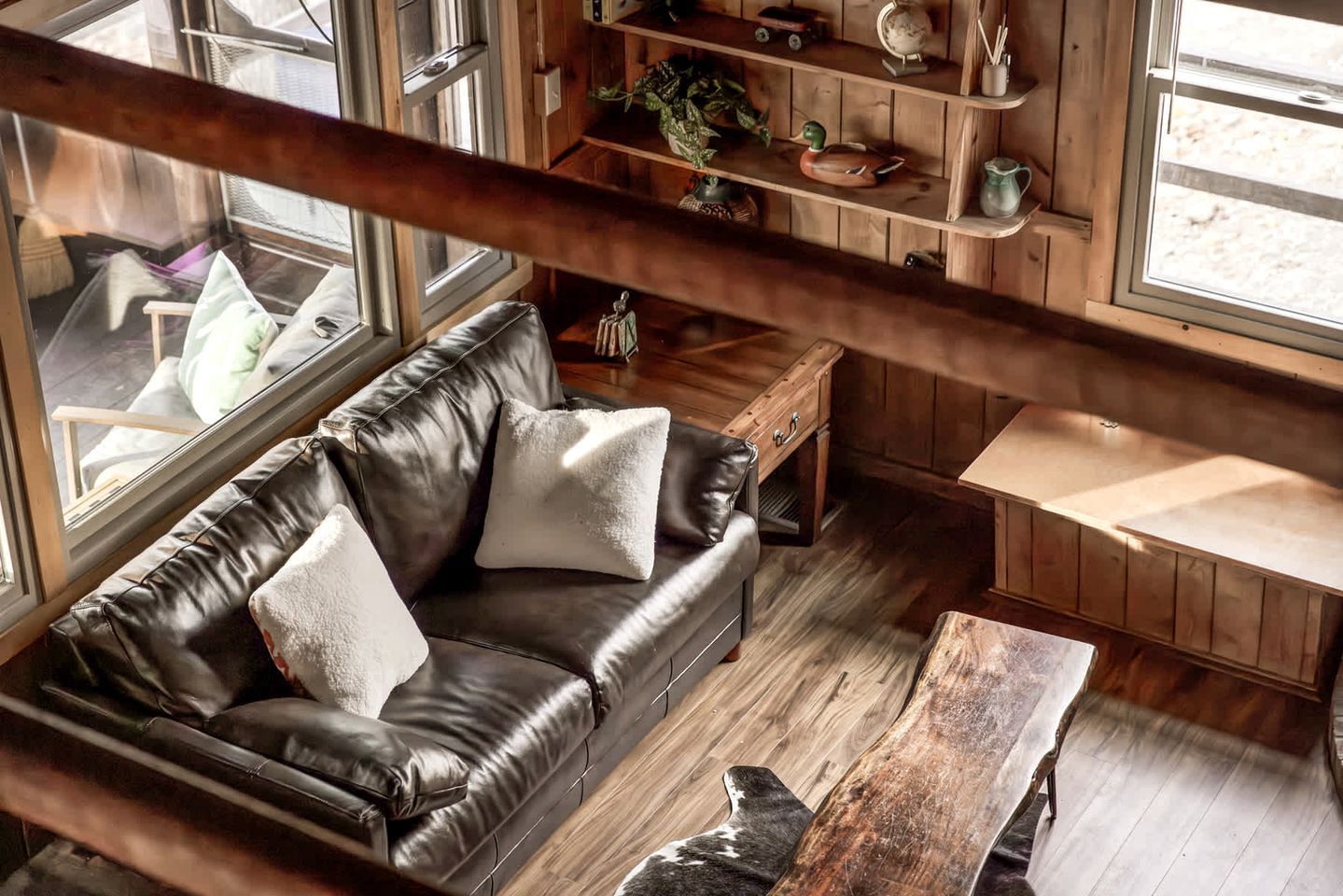 A cozy room with a leather sofa, wooden shelves, and a view through large windows.