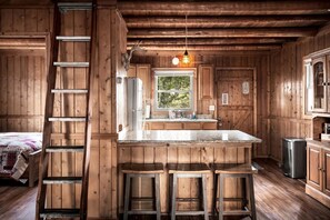 A wooden cabin interior with a central island, bar stools, a ladder, and a window with a view of greenery.