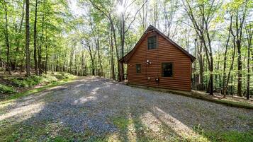A wooden cabin with a gravel driveway, surrounded by a forest.