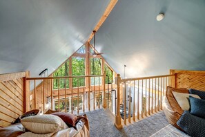 A cozy attic bedroom with a wooden ceiling, a balcony with a railing, and a view of the forest.