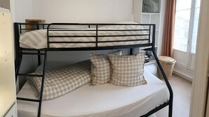 A bunk bed with a white mattress and checkered pillows, a white cabinet, and a window with curtains. - Bright and renovated, City center (Vitry-le-François)