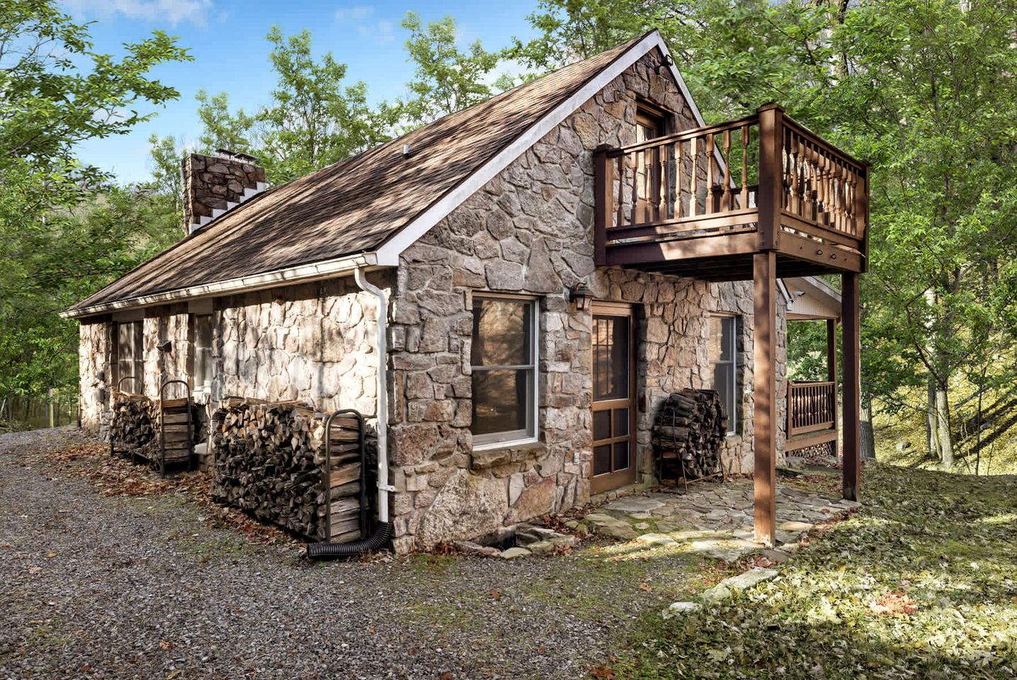 A rustic stone cabin with a wooden balcony, a stone chimney, and a gravel driveway.