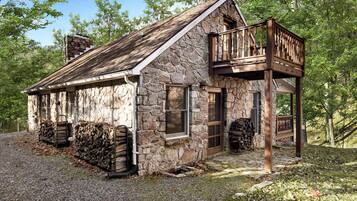 A rustic stone cabin with a wooden balcony, a stone chimney, and a gravel driveway.