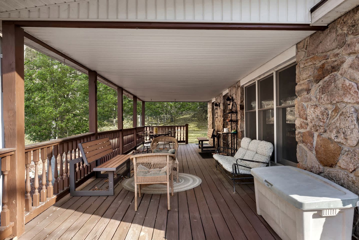 Une véranda couverte avec un plancher en bois, un mur en pierre et du mobilier d’extérieur comprenant un banc et des chaises.