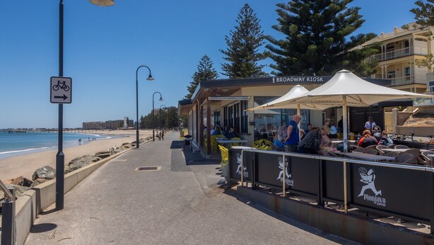 A coastal walkway with a bike path sign, a kiosk named 'Broadway Kiosk', and outdoor seating with people.
