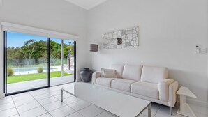 A modern living room with a white sofa, a coffee table, and a floor lamp. There is a large window with a view of a garden and a swimming pool. - Beachfront 3-bed With Private Pool & Fireplace (Sunshine Coast)