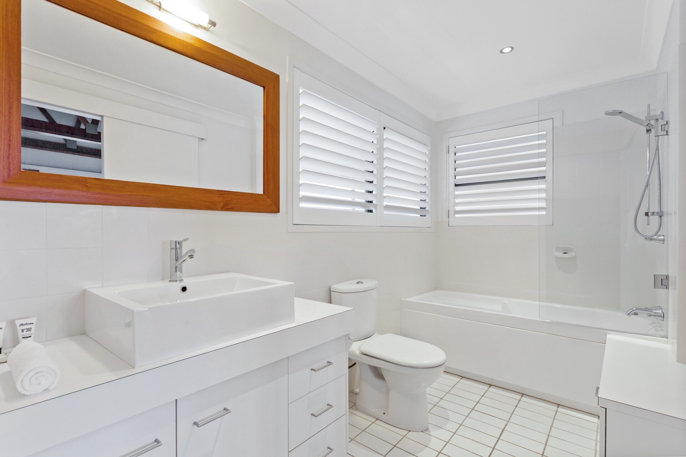 Une salle de bain moderne avec un lavabo, des toilettes et une baignoire blancs, ainsi qu’un grand miroir doté d’un cadre en bois.