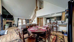 Un vaste espace de vie comprenant un coin repas avec une table pour quatre personnes, une cheminée et une cuisine en arrière-plan.